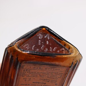 K&ouml;nnte beinhalten: Nahaufnahme einer Vintage-Flasche aus bernsteinfarbenem Glas mit dreieckiger Oberseite. Die Oberseite hat einen gepr&auml;gten Text, und ein Etikett mit Text ist sichtbar. Die Flasche hat eine rechteckige Form mit vertikalen Rippen.