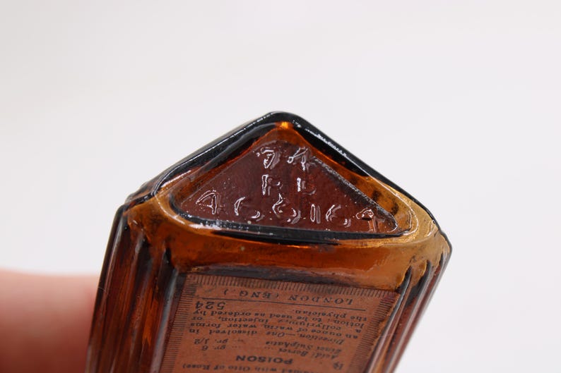 K&ouml;nnte beinhalten: Nahaufnahme einer Vintage-Flasche aus bernsteinfarbenem Glas mit dreieckiger Oberseite. Die Oberseite hat einen gepr&auml;gten Text, und ein Etikett mit Text ist sichtbar. Die Flasche hat eine rechteckige Form mit vertikalen Rippen.
