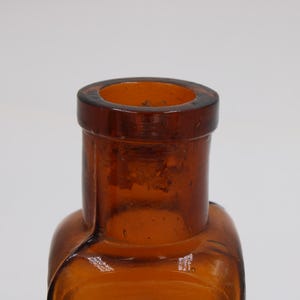 May include: Close-up of a vintage amber glass bottle. The bottle has a rectangular body with rounded edges and a cylindrical neck. The glass has a slightly textured surface and a dark amber color.