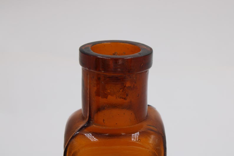 May include: Close-up of a vintage amber glass bottle. The bottle has a rectangular body with rounded edges and a cylindrical neck. The glass has a slightly textured surface and a dark amber color.
