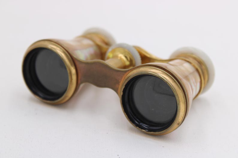 May include: Antique opera glasses with a brass frame and mother-of-pearl accents. The lenses are black, and the binoculars have a handle for easy grip. The design is vintage and elegant.
