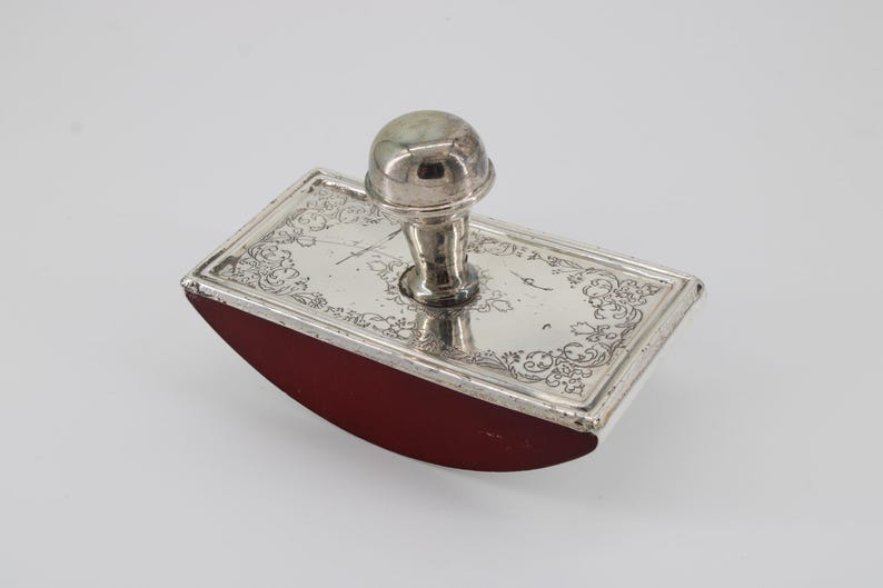May include: A vintage silver ink blotter with a rectangular base and a rounded, red blotting surface. The silver top is decorated with ornate floral patterns and a central handle. A classic desk accessory.