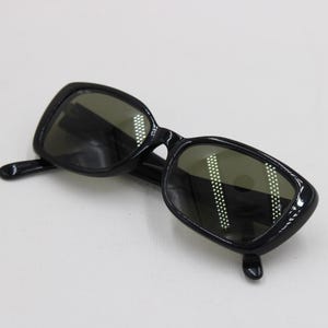 Puede incluir: Gafas de sol negras con montura rectangular y lentes oscuras. Las gafas de sol tienen un acabado brillante y est&aacute;n sobre un fondo blanco. Las lentes reflejan la luz.