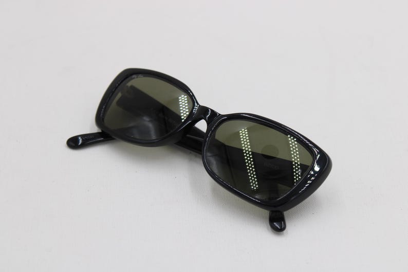 Puede incluir: Gafas de sol negras con montura rectangular y lentes oscuras. Las gafas de sol tienen un acabado brillante y est&aacute;n sobre un fondo blanco. Las lentes reflejan la luz.
