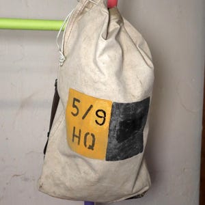 May include: A canvas bag with a drawstring closure, hanging from colorful poles. The bag has a yellow patch with the numbers "5/9" and the letters "HQ" in black. A black square is next to the text.