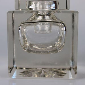 May include: Clear glass perfume bottle with a square base and a rounded, bulbous center. The bottle has a decorative, starburst pattern on the bottom. The top of the bottle has a small, cylindrical stopper.