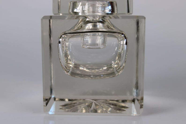May include: Clear glass perfume bottle with a square base and a rounded, bulbous center. The bottle has a decorative, starburst pattern on the bottom. The top of the bottle has a small, cylindrical stopper.