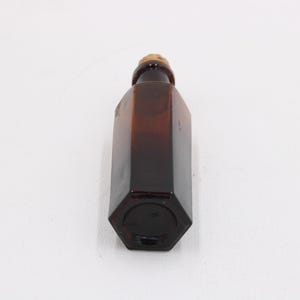 Puede incluir: Una botella de vidrio hexagonal marr&oacute;n con un tap&oacute;n de corcho. La botella es de color &aacute;mbar oscuro y tiene forma geom&eacute;trica. El corcho es de color marr&oacute;n claro. La botella est&aacute; vac&iacute;a y sobre una superficie blanca.