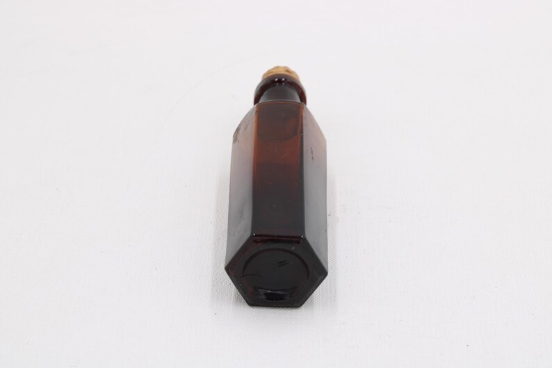 Puede incluir: Una botella de vidrio hexagonal marr&oacute;n con un tap&oacute;n de corcho. La botella es de color &aacute;mbar oscuro y tiene forma geom&eacute;trica. El corcho es de color marr&oacute;n claro. La botella est&aacute; vac&iacute;a y sobre una superficie blanca.