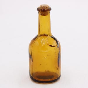 May include: Amber-colored glass bottle with a cork stopper. The bottle has a rounded body and a long, slender neck. The cork is a light brown color. The bottle is empty and sits on a white background.