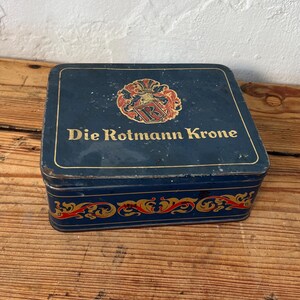May include: A rectangular, vintage blue tin with the words "Die Rotmann Krone" in gold lettering. The tin features a decorative crest and ornate gold and red scrollwork. The tin sits on a wooden surface.
