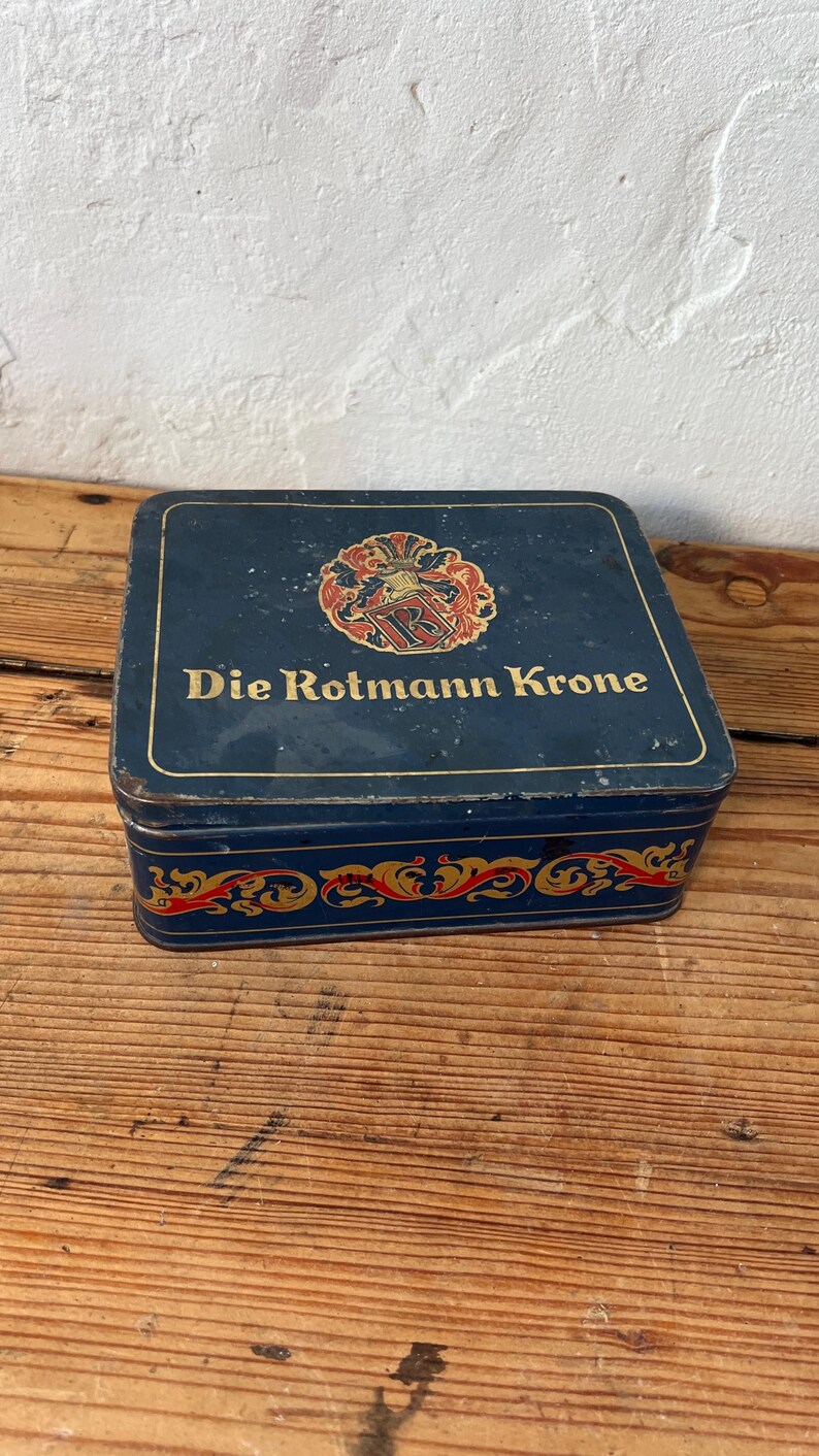 May include: A rectangular, vintage blue tin with the words "Die Rotmann Krone" in gold lettering. The tin features a decorative crest and ornate gold and red scrollwork. The tin sits on a wooden surface.