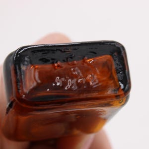 May include: Close-up of the bottom of a small, rectangular amber glass bottle. The bottle has a dark brown, glossy rim and the base has embossed text. The bottle is held by a person.