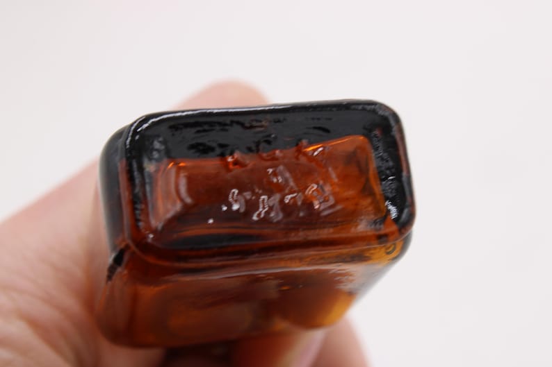 May include: Close-up of the bottom of a small, rectangular amber glass bottle. The bottle has a dark brown, glossy rim and the base has embossed text. The bottle is held by a person.