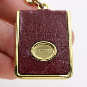 May include: A rectangular keychain with a burgundy leather surface and gold-colored trim. The center features a gold oval with a stylized design. The keychain is attached to a gold chain.