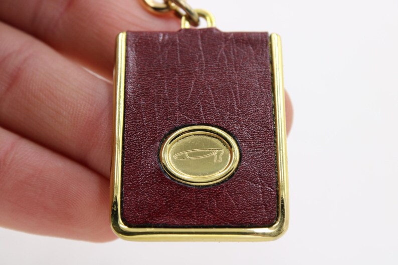 May include: A rectangular keychain with a burgundy leather surface and gold-colored trim. The center features a gold oval with a stylized design. The keychain is attached to a gold chain.