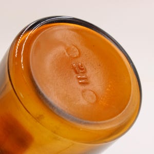 May include: Close-up of the bottom of a cylindrical amber glass bottle. The base is a lighter shade of amber with embossed text and circular markings. The bottle's sides are a darker amber color.