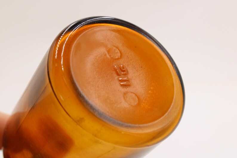 May include: Close-up of the bottom of a cylindrical amber glass bottle. The base is a lighter shade of amber with embossed text and circular markings. The bottle's sides are a darker amber color.