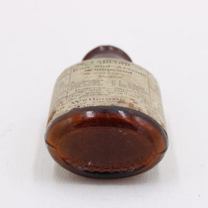 May include: A vintage brown glass bottle with a label wrapped around it. The label has faded text. The bottle is lying on its side, showcasing its rounded base and the label's details. The bottle appears to be an antique medicine bottle.