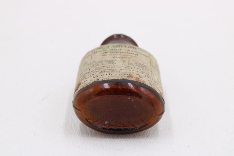 May include: A vintage brown glass bottle with a label wrapped around it. The label has faded text. The bottle is lying on its side, showcasing its rounded base and the label's details. The bottle appears to be an antique medicine bottle.