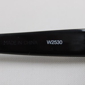 Puede incluir: Primer plano de la patilla de unas gafas de sol negras. El texto "MADE IN CHINA" y "W2530" est&aacute;n impresos en blanco en la patilla. La bisagra es visible.