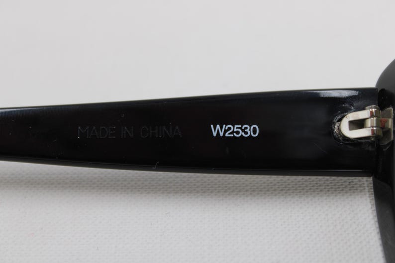 Puede incluir: Primer plano de la patilla de unas gafas de sol negras. El texto "MADE IN CHINA" y "W2530" est&aacute;n impresos en blanco en la patilla. La bisagra es visible.