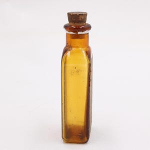 May include: An amber-coloured antique glass bottle with a cork stopper. The rectangular bottle has rounded corners and a slightly textured surface. The cork is a dark brown colour. The bottle is empty and sits against a white background.