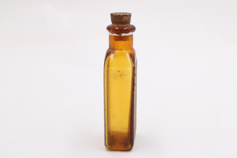 May include: An amber-coloured antique glass bottle with a cork stopper. The rectangular bottle has rounded corners and a slightly textured surface. The cork is a dark brown colour. The bottle is empty and sits against a white background.