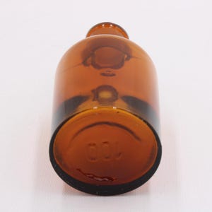 May include: A brown glass bottle with a rounded body and a narrow neck. The bottom of the bottle has the number "100" embossed. The bottle is made of amber-colored glass.