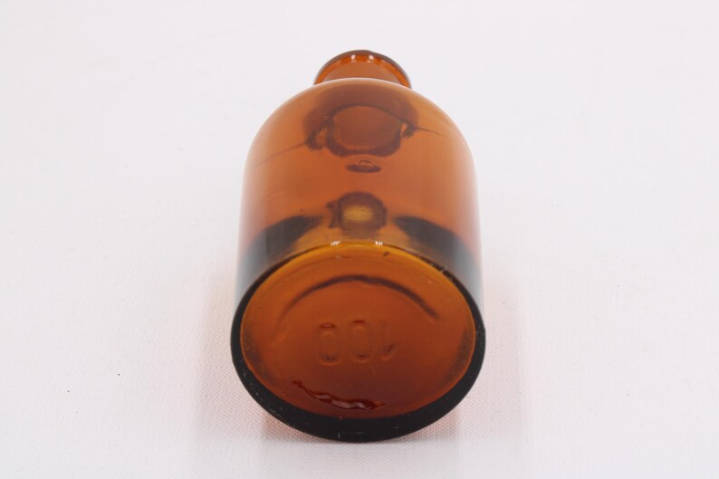 May include: A brown glass bottle with a rounded body and a narrow neck. The bottom of the bottle has the number "100" embossed. The bottle is made of amber-colored glass.