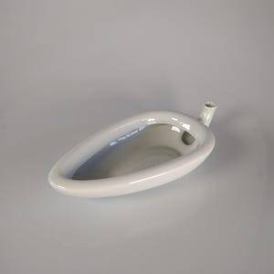 May include: A white, ceramic urinal with a rounded, elongated shape. It features a small, cylindrical spout extending from the side. The urinal is clean and appears unused, set against a plain, light gray background.
