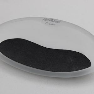 K&ouml;nnte beinhalten: Oval geformte Schale aus mattiertem Glas mit einem schwarzen, nierenf&ouml;rmigen Einsatz. Der Text "Apollo Optik 25 Jahre" ist grau auf der Oberseite aufgedruckt. Die Schale dient wahrscheinlich zum Halten kleiner Gegenst&auml;nde.