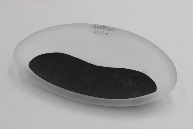 K&ouml;nnte beinhalten: Oval geformte Schale aus mattiertem Glas mit einem schwarzen, nierenf&ouml;rmigen Einsatz. Der Text "Apollo Optik 25 Jahre" ist grau auf der Oberseite aufgedruckt. Die Schale dient wahrscheinlich zum Halten kleiner Gegenst&auml;nde.