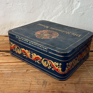 May include: A rectangular, vintage blue tin with gold and red decorative scrollwork. The tin has the words "Die Rotmann Krone" and "FR. ROTMANN BURGSTEINFURT" printed on it. The tin is sitting on a wooden surface.