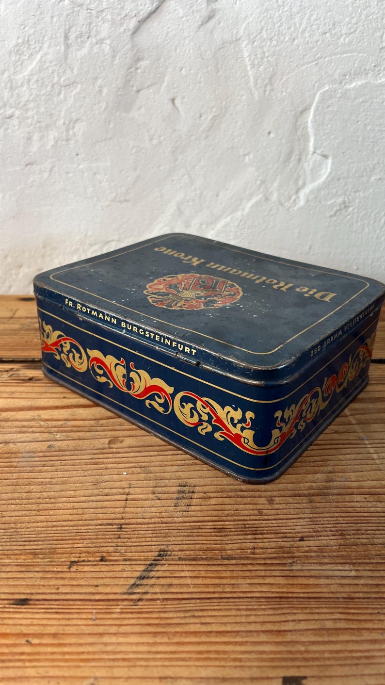May include: A rectangular, vintage blue tin with gold and red decorative scrollwork. The tin has the words "Die Rotmann Krone" and "FR. ROTMANN BURGSTEINFURT" printed on it. The tin is sitting on a wooden surface.