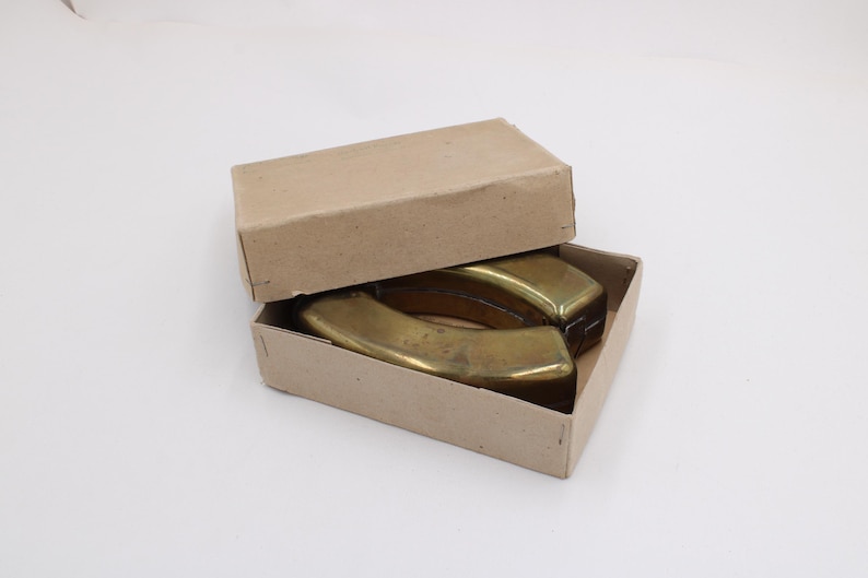 May include: Two brass-colored, curved objects are inside a partially open, rectangular cardboard box. The box is light brown. The objects have a rounded shape and appear to be made of metal. The box is on a white surface.