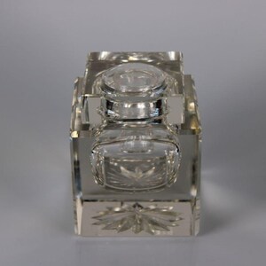 May include: A clear, rectangular crystal inkwell with a lid and a small, square ink pot. The inkwell features intricate starburst and floral patterns on its base and sides. The design is symmetrical and elegant.