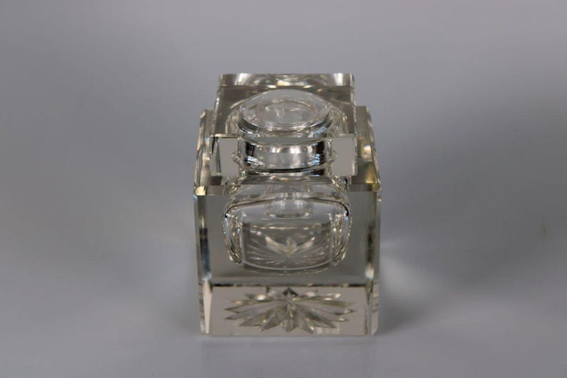 May include: A clear, rectangular crystal inkwell with a lid and a small, square ink pot. The inkwell features intricate starburst and floral patterns on its base and sides. The design is symmetrical and elegant.