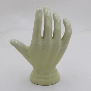 May include: A pale green ceramic hand sculpture with a rounded base. The hand is open, with fingers slightly curved. The sculpture is smooth and glossy, and the fingers are separated.