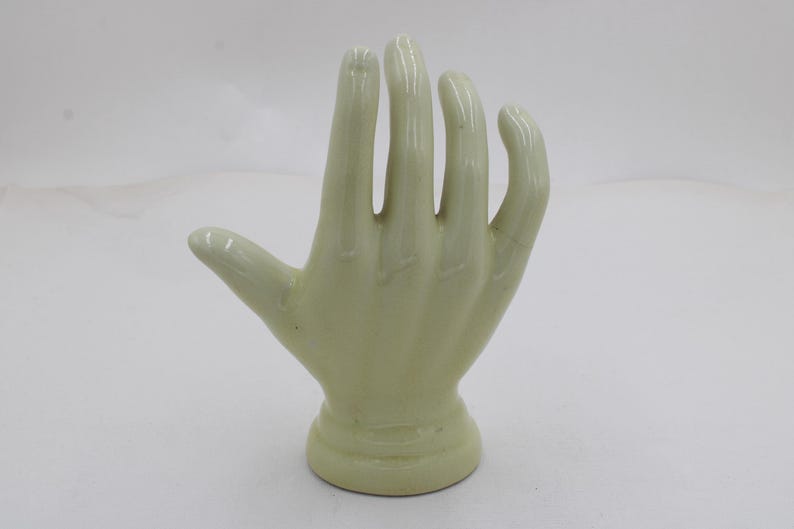 May include: A pale green ceramic hand sculpture with a rounded base. The hand is open, with fingers slightly curved. The sculpture is smooth and glossy, and the fingers are separated.
