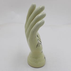 May include: A light green ceramic hand sculpture. The hand is in a curved position, with the fingers slightly bent. Black line art decorates the base of the hand. The sculpture is on a white background.