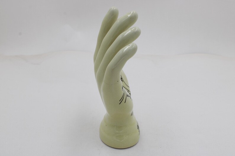 May include: A light green ceramic hand sculpture. The hand is in a curved position, with the fingers slightly bent. Black line art decorates the base of the hand. The sculpture is on a white background.