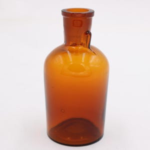 May include: Amber-colored glass bottle with a rounded body and a narrow neck. The bottle has a small handle-like protrusion near the neck. The glass has a slight amber hue, and the bottle is empty.