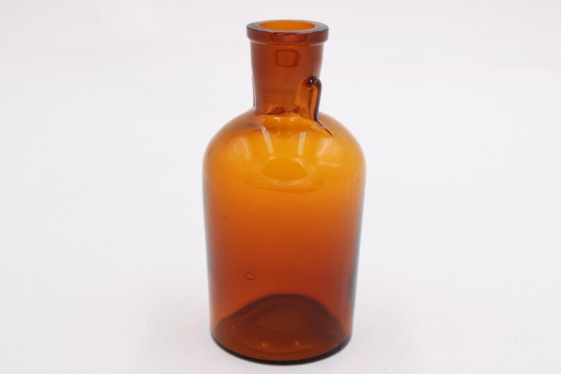 May include: Amber-colored glass bottle with a rounded body and a narrow neck. The bottle has a small handle-like protrusion near the neck. The glass has a slight amber hue, and the bottle is empty.