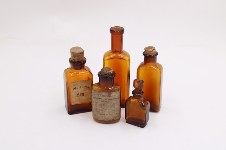 May include: Five antique amber glass bottles with cork stoppers. Two bottles have labels that read "METHYL SAL" and "CARBOLIC POISONING COMPOUND". The bottles vary in shape and size, with a square, rectangular, and cylindrical design.