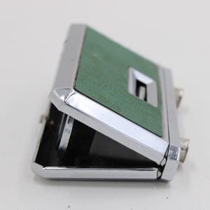 May include: A vintage pair of opera glasses with a green textured top and a silver-colored metal frame. The binoculars are partially open, revealing the lenses. The design is compact and classic.
