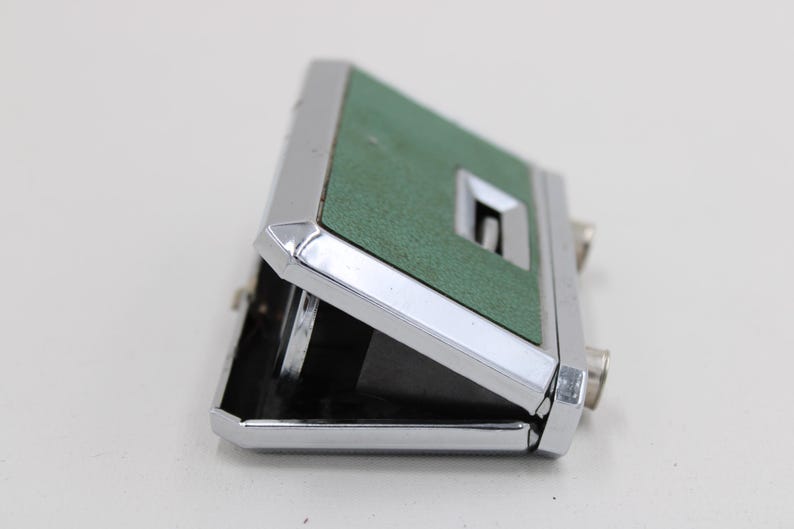 May include: A vintage pair of opera glasses with a green textured top and a silver-colored metal frame. The binoculars are partially open, revealing the lenses. The design is compact and classic.