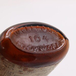 May include: Close-up of the base of a brown glass bottle. The text "WELLCOME" is arched across the top, with "104" below it. "CHEM. WORKS" is arched across the bottom. The bottle has a vintage appearance.