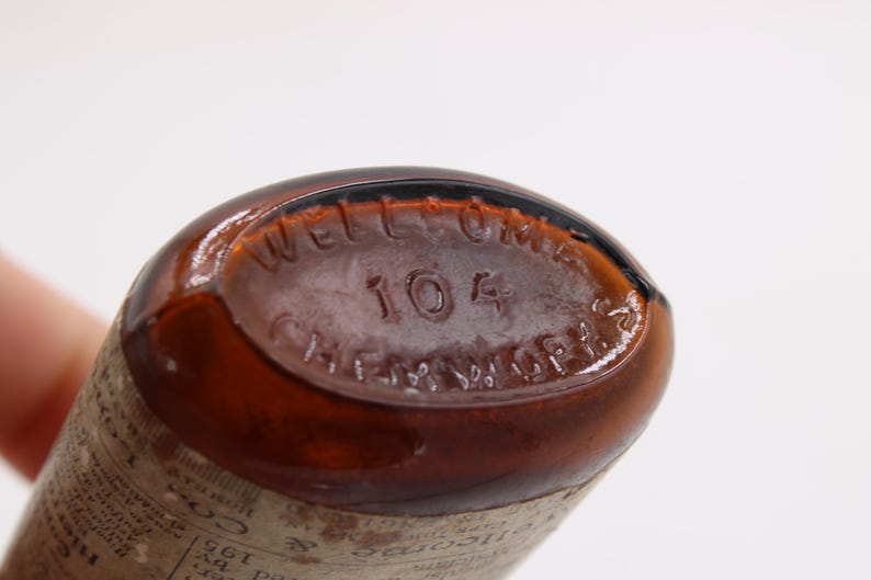 May include: Close-up of the base of a brown glass bottle. The text "WELLCOME" is arched across the top, with "104" below it. "CHEM. WORKS" is arched across the bottom. The bottle has a vintage appearance.