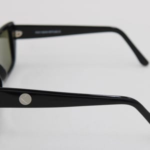 Puede incluir: Gafas de sol negras con montura rectangular y lentes oscuras. Las patillas son elegantes y presentan un emblema circular plateado. Las gafas de sol se encuentran sobre un fondo blanco liso, mostrando su dise&ntilde;o moderno.